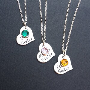 May include: Three silver necklaces with heart-shaped pendants. Each pendant has a different colored gemstone and the words "Big Sister", "Middle Sister", and "Little Sister" engraved on them.