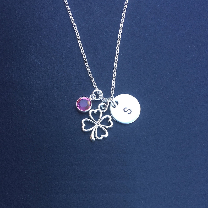May include: A silver necklace with a four-leaf clover charm, a pink crystal, and a silver disc with the letter 'S' engraved on it.