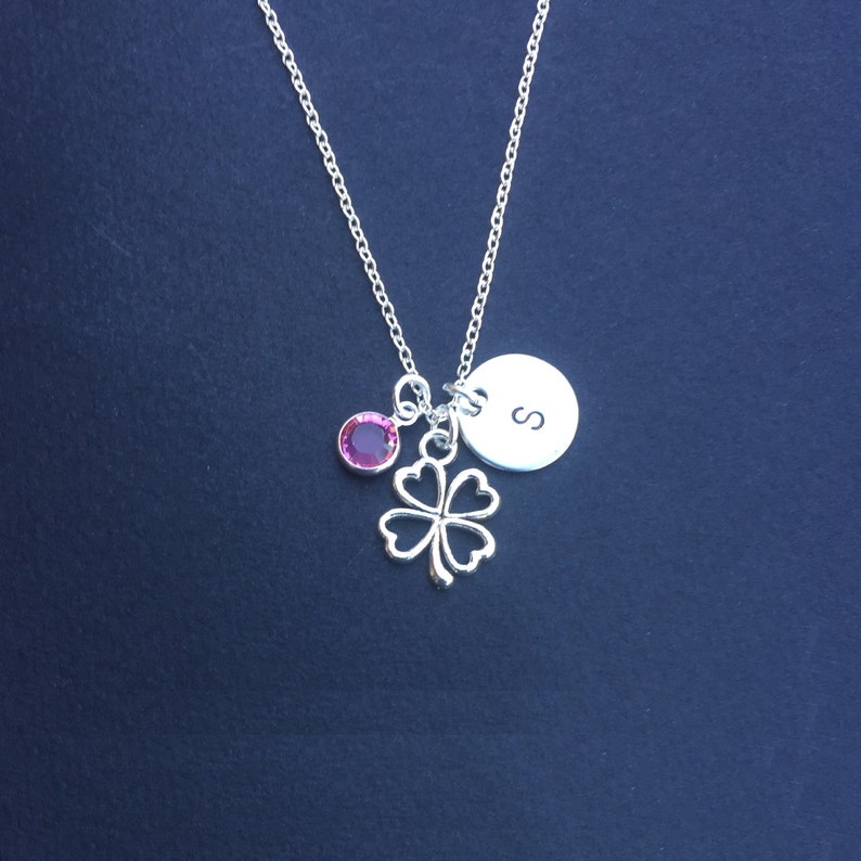 May include: Silver necklace with a four-leaf clover charm, a small pink crystal, and a round charm with the letter 'S' engraved on it.