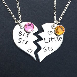 May include: Two silver necklaces with heart-shaped pendants that say "Big Sis" and "Little Sis". The pendants are broken in half and each has a small crystal, one pink and one orange.