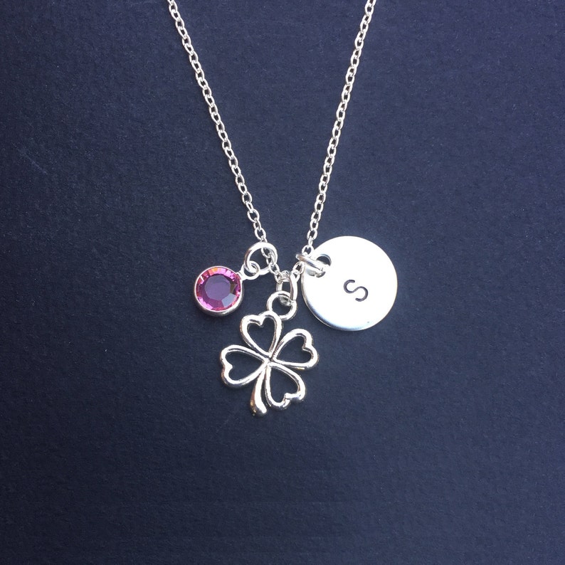 May include: A silver necklace with a four-leaf clover charm, a pink crystal, and a silver disc with the letter "S" engraved on it.