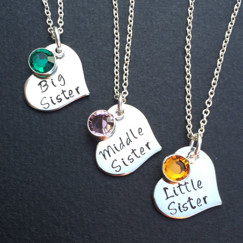 May include: Three silver heart-shaped necklaces with different colored gemstones. The necklaces say "Big Sister", "Middle Sister", and "Little Sister".