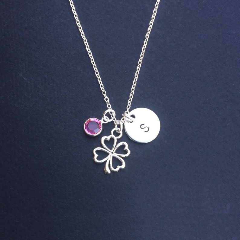 May include: Silver necklace with a four-leaf clover charm, a pink crystal, and a round charm with the letter "S" engraved on it.