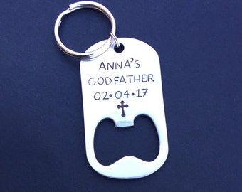 May include: A silver bottle opener keychain with the text "ANNA'S GODFATHER 02.04.17" and a cross symbol. The keychain has a ring at the top and a bottle opener at the bottom. The background is a dark blue.