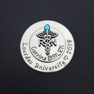 May include: Silver round charm with a blue gem on top. The charm features a caduceus medical symbol with the letters "RN" and the text "Corrine BSN. RN 2018 Lourdes University".