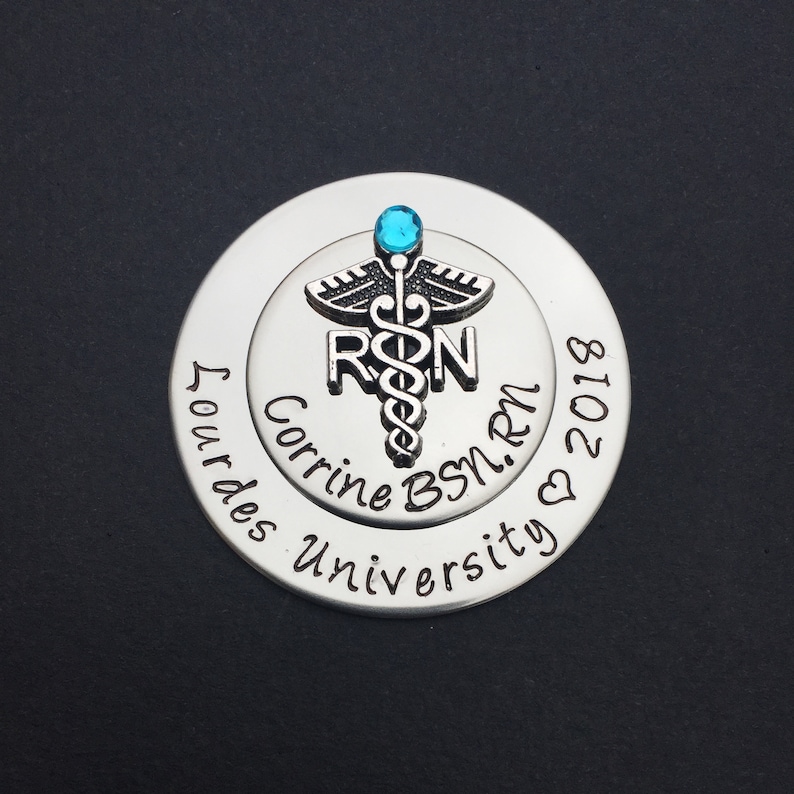 May include: Silver round charm with a blue gem on top. The charm features a caduceus medical symbol with the letters "RN" and the text "Corrine BSN. RN 2018 Lourdes University".