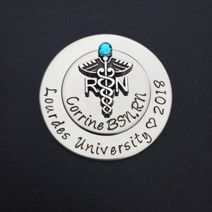 May include: Silver round charm with a blue rhinestone and a medical caduceus symbol. The charm is engraved with the text "Corrine BSN, RN 2018" and "Our Lady of Lourdes University".