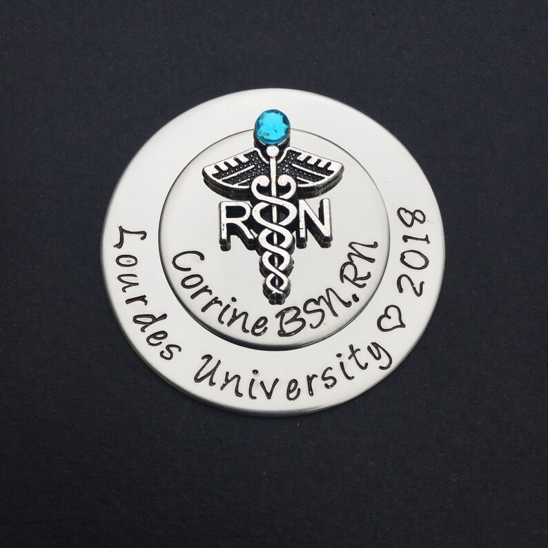 May include: Silver round charm with a blue rhinestone and a medical caduceus symbol. The charm is engraved with the text "Corrine BSN, RN 2018" and "Our Lady of Lourdes University".