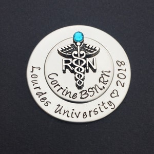 May include: Silver round charm with a blue gem on top of a caduceus symbol with the letters RN. The charm also has the text "Corrine BSN, RN" and "Our Lady University" with a heart and the year 2018.