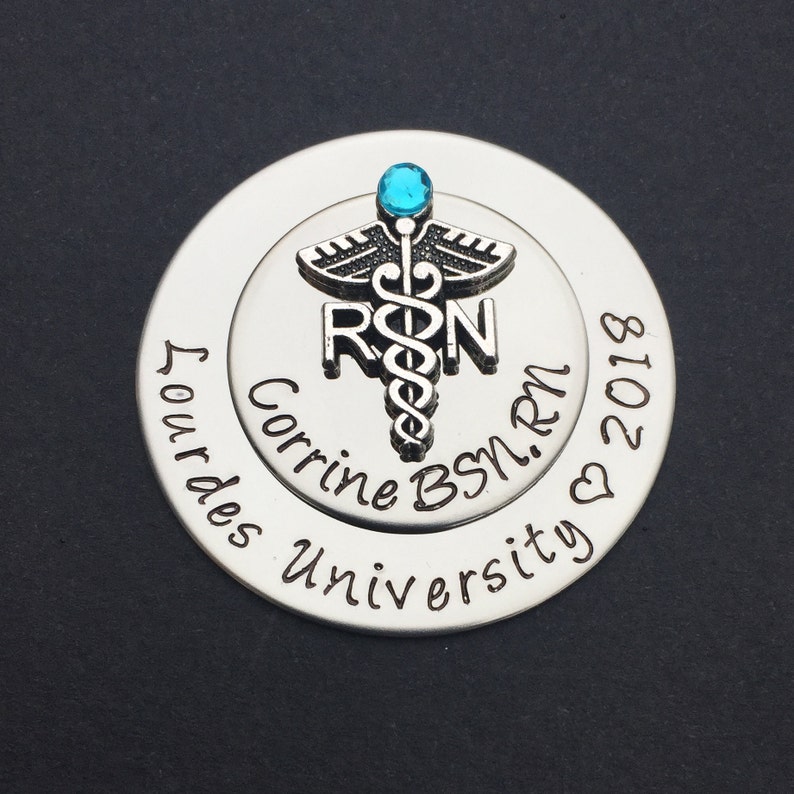May include: Silver round charm with a blue gem on top of a caduceus symbol with the letters RN. The charm also has the text "Corrine BSN, RN" and "Our Lady University" with a heart and the year 2018.