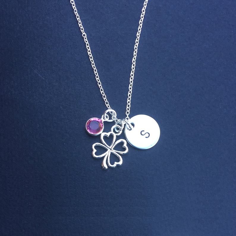 May include: Silver necklace with a four-leaf clover charm, a pink gemstone, and a round charm with the letter "S" engraved on it.