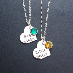 May include: Two silver necklaces with heart pendants. One pendant says "Big Sister" with a green gemstone, the other says "Little Sister" with a yellow gemstone. Both pendants are on delicate silver chains.
