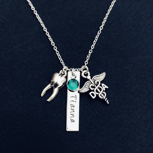 May include: A silver necklace with a tooth charm, a green gemstone, a personalized bar with the name "Tianna" engraved on it, and a caduceus charm.