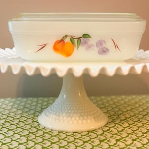 May include: A white rectangular dish with a lid, decorated with orange and purple fruit illustrations, sits on a white, ruffled-edge cake stand. The stand has a pedestal base with a textured, dotted pattern. The background is a patterned green and white tablecloth.