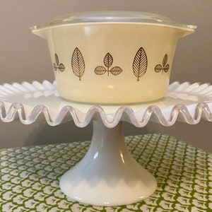 May include: A white ceramic cake stand with a scalloped edge and a white pedestal base. A yellow casserole dish with a brown leaf design is on top of the stand.