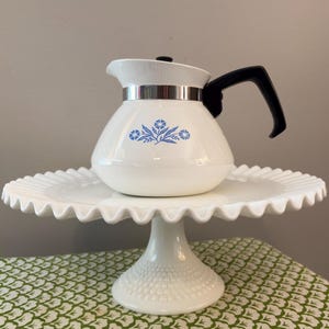 May include: A white teapot with a blue floral pattern, a black handle, and a silver-tone metal band around the neck. The teapot sits on a white milk glass cake stand featuring a ruffled edge and a hobnail textured base.
