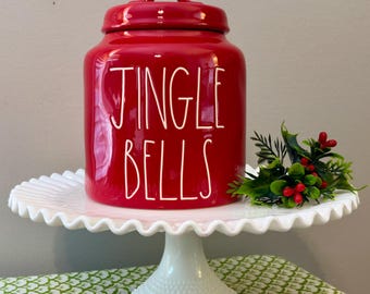 Puede incluir: Un tarro de cerámica rojo brillante con tapa, que presenta el texto blanco JINGLE BELLS en una fuente manuscrita alta y delgada. El tarro está colocado sobre un soporte para pasteles blanco festoneado junto a una ramita de acebo verde.