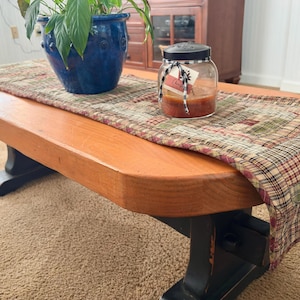 May include: A rectangular wooden coffee table with a natural wood top and black legs, featuring a plaid fabric table runner in shades of red, green, and tan. A blue ceramic planter with a green plant and a glass candle jar sit on the runner.