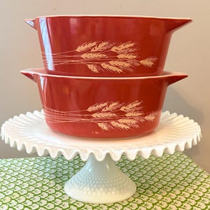 May include: Two red casserole dishes with wheat stalk designs are stacked on a white, ruffled-edge cake stand. The dishes have handles and a white rim. The cake stand has a textured base and a scalloped edge.