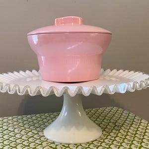 May include: A pink ceramic serving dish with a lid sits on a white cake stand with a scalloped edge. The stand is on a green and white patterned tablecloth.