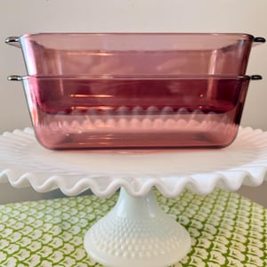 May include: Two rectangular, pink glass baking dishes with silver handles are stacked on a white, ruffled-edge cake stand. The stand has a pedestal base and sits on a green and white patterned tablecloth.