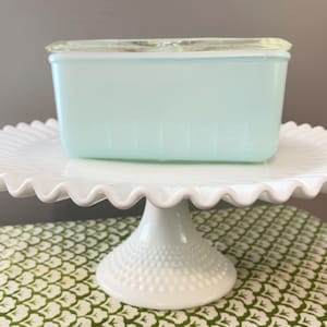 May include: A white, ruffled-edge cake stand with a light blue rectangular container with a clear lid. The cake stand has a pedestal base with a textured, dotted pattern. The container appears to be made of glass.