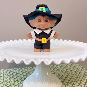 May include: A small troll doll with teal hair wearing a black pilgrim-style hat with a blue ribbon and yellow buckle, a black outfit with a white collar, and an orange belt with a yellow buckle, standing on a white ruffled cake stand.