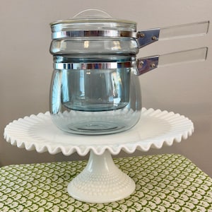 May include: A vintage blue glass double boiler with clear handles and a lid, set on a white ruffled cake stand. The stand has a pedestal base and sits on a green and white patterned surface.