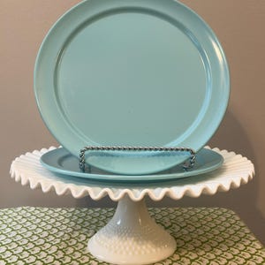 May include: Two smooth, solid light blue plates are stacked on a white milk glass cake stand. The stand features a ruffled edge and a textured hobnail base. A silver-toned, twisted metal wire stand holds the top plate upright.
