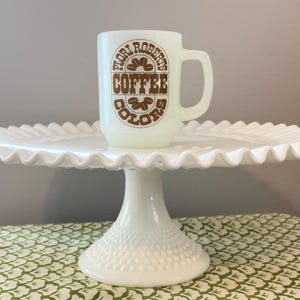 May include: A white ceramic mug with the text "Flori Roberts Coffee Colors" in brown lettering, featuring coffee bean graphics. The mug sits on a white, ruffled-edge cake stand with a pedestal base.