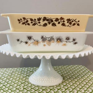 May include: Two rectangular glass baking dishes stacked on a white milk glass cake stand. The top dish is pale yellow with a brown leaf pattern. The bottom dish is white with a brown and gold pinecone and needle design. The stand has a ruffled edge.