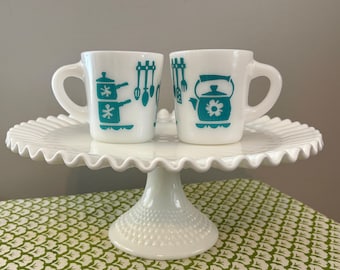 May include: Two white ceramic mugs with teal kitchen utensil designs, including pots, spoons, and a teapot with a flower. The mugs sit on a white, ruffled-edge cake stand. The stand is on a green and white patterned tablecloth.