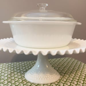 May include: A white, vintage casserole dish with a clear glass lid, resting on a white, scalloped-edge cake stand. The dish has a textured design. The cake stand has a pedestal base with a beaded pattern. The background is a neutral color.