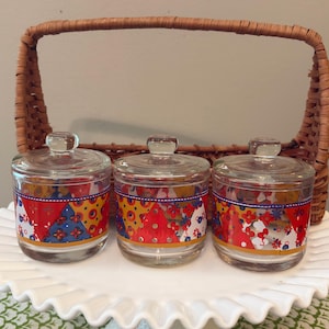 May include: Three clear glass jars with matching lids, each featuring a colorful band of red, yellow, and blue geometric and floral patterns. The jars are arranged in a row in front of a brown woven wicker basket with a tall arched handle.