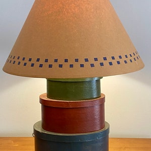 May include: A unique table lamp with a tan lampshade featuring a blue square pattern. The lamp base is composed of stacked, painted round boxes in green, red, and blue. The lamp sits on a wooden surface.