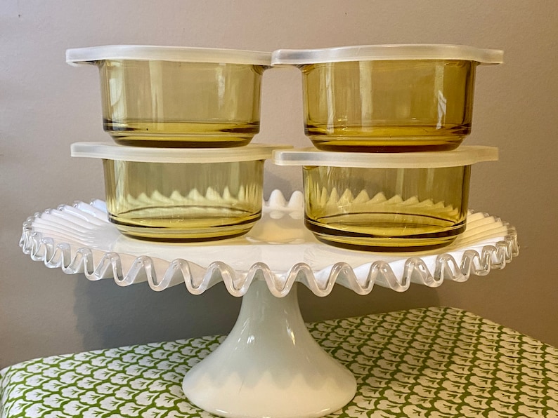May include: Four amber glass storage containers with white lids on a white cake stand. The containers are stacked in pairs.