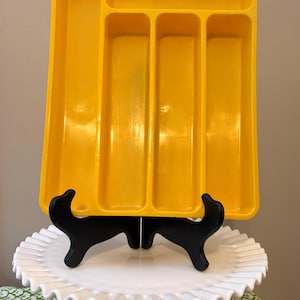 May include: A bright yellow plastic utensil tray with multiple compartments. The tray is displayed on a black stand atop a white, ruffled-edge cake stand. The background is a neutral color.