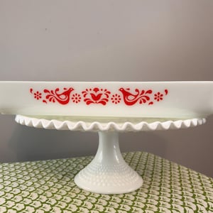 May include: A rectangular white glass baking dish featuring a red printed pattern of two stylized birds facing each other, surrounded by floral and leaf motifs. The dish has integrated side handles and sits atop a white pedestal cake stand.