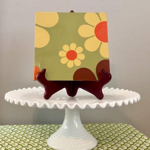 May include: A square, floral-patterned artwork in shades of yellow, orange, and brown on a green background, displayed on a dark red easel. The easel rests on a white, ruffled-edge cake stand, set atop a green and white patterned tablecloth.