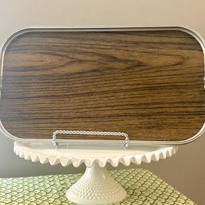 May include: A rectangular serving tray with a wood grain pattern and silver-colored metal trim and handles. The tray is displayed on a white, scalloped-edge cake stand, atop a green and white patterned tablecloth.