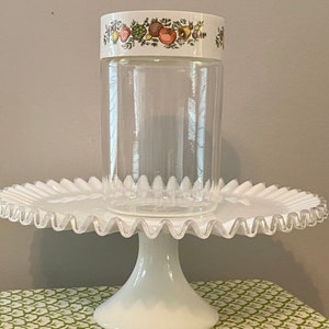 May include: A clear glass jar with a white lid featuring a colorful fruit and vegetable design. The jar sits on a white, ruffled-edge cake stand. The stand has a pedestal base.