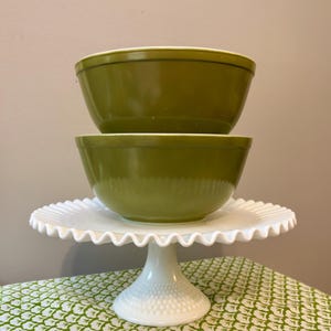 May include: Two stacked olive green mixing bowls with smooth, rounded sides resting on a white milk glass cake stand. The cake stand features a ruffled edge and a textured, hobnail-style base, providing a stable platform for the two bowls.