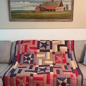 May include: A rectangular quilt featuring a patchwork design of red, navy blue, and cream fabric squares. The pattern incorporates multiple star motifs set against a background of striped and floral prints, creating a geometric, multi-colored textile.