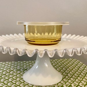 May include: A white cake stand with a scalloped edge and a clear glass bowl with a lid on top. The bowl is amber in color.