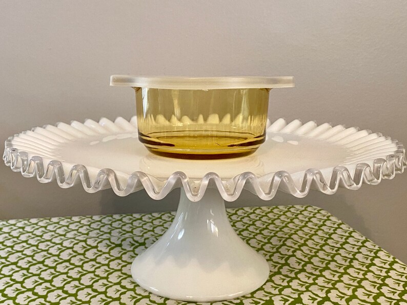 May include: A white cake stand with a scalloped edge and a clear glass bowl with a lid on top. The bowl is amber in color.