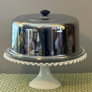 May include: A silver-colored cake stand with a domed lid and a black knob. The stand has a white, scalloped edge and a white pedestal base. The reflective surface of the lid mirrors the surroundings.