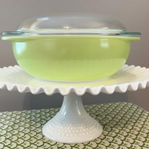May include: A lime green Pyrex bowl with a clear glass lid sits atop a white, ruffled-edge cake stand. The stand has a pedestal base with a beaded texture. The bowl is a vibrant, solid color, and the stand adds a touch of elegance.