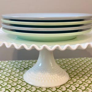 May include: A white, ruffled-edge cake stand with a stack of pastel-colored plates. The plates are light blue, green, and gray. The stand has a pedestal base with a textured, dotted pattern.