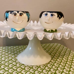 May include: Two ceramic cups with painted faces, one with blue hair and a blue and green outfit, the other with black hair and a green outfit, on a white cake stand with a scalloped edge.
