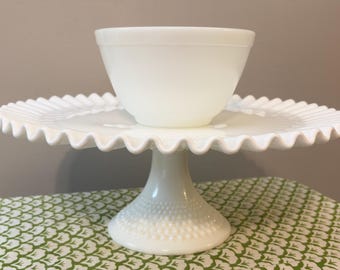 May include: A white milk glass cake stand with a ruffled edge and a pedestal base featuring a hobnail texture, topped with a matching white milk glass bowl. The cake stand has a wide, flat plate surface and a sturdy, circular base.